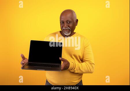 Porträt eines glücklichen älteren schwarzen Mannes, der einen Laptop mit leerem Bildschirm vorführt, auf orangefarbenem Studiohintergrund posiert und ein Modell vorführt Stockfoto
