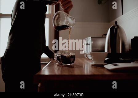 Der junge, tausendjährige Hipster-Mischling gießt frisch gemahlenen und gebrühten Kaffee in seiner Küche zu Hause in Glas-Becher Stockfoto