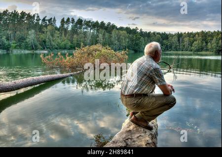 Im letzten Licht der untergehenden Sonne sitzt ein alter Mann auf einem Baumstamm auf einem einsamen See und genießt die Stille und Stille der Abendstunde in der Natur. Stockfoto