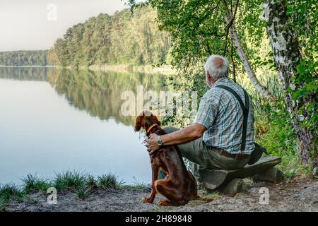 Am frühen Morgen sitzt ein alter Mann mit seinem jungen irischen Setter Pointer auf der kleinen Bank am See und genießt die Stille des Anfangstages. Stockfoto