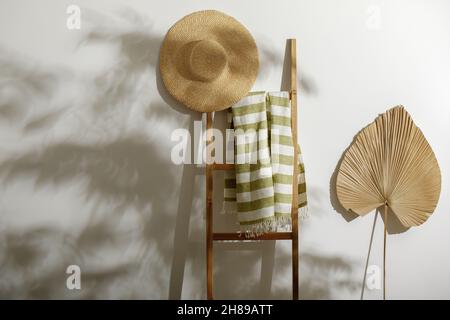 Strandtuch aus Bio-Leinen auf Holzleiter, Strohhut und getrocknetes Palmblatt an der weißen Wand mit Schatten von Baumblättern Stockfoto