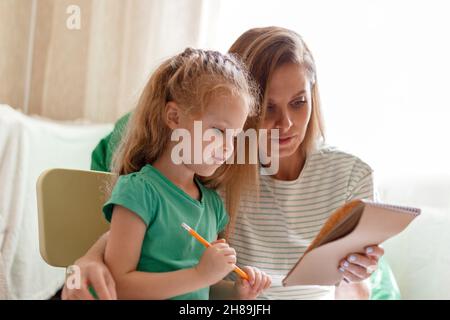 Kaukasische blonde intelligente Kind Kind 5 Jahre Hausaufgaben machen Lernen, mit Mama 30s zusammen zu schreiben. Mutter hilft Vorschulmädchen Tochter Hausaufgaben zu machen Stockfoto