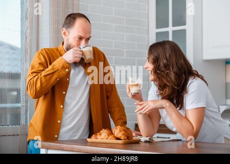 Ein paar lächelnde junge Leute, Mann und Frau, die zu Hause in der Küche frühstücken. Das Ehepaar trank Kaffee und aß Croissants in ihrem Stockfoto