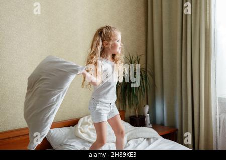 Fröhliches kleines Kind Mädchen mit Spaß im Schlafzimmer spielen mit Kissen. Vorschule Tochter kaukasisch blonde Mädchen spielen in Kissen Kissen Kampf im Schlafzimmer Stockfoto