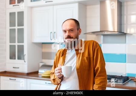 Portrait junger glücklicher Mann, der Kaffee oder Tee in der Küche zu Hause trinkt. Glatze bärtig 30s Mann genießt morgendliche Routine in Küche Wohnung. Stockfoto