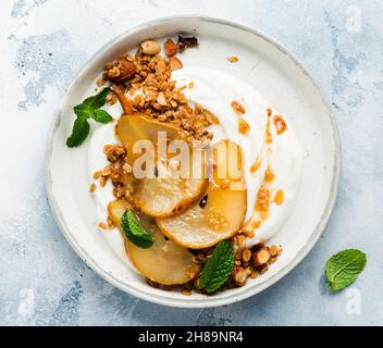 Griechischer Joghurt mit karamellisierter Birne, Müsli, Nüssen und geschmolzenem Zucker für ein gesundes Frühstück auf einer grauen Keramikplatte. Rustikaler Stil. Draufsicht. Stockfoto