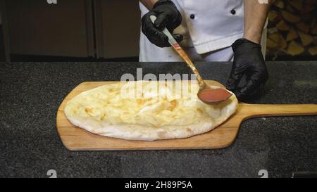 Nahaufnahme des Küchenchefs bereitet köstliche italienische Pizza im Restaurant zu. Rahmen. Der Koch gießt Tomatensauce auf gebackene runde Pizzabasis. Pizza im Ofen auf Woo gekocht Stockfoto