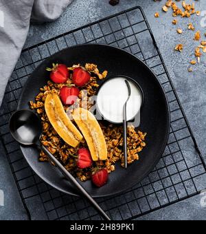 Haferflockengranola mit Joghurt, frischen Erdbeeren und Banane, Chiasamen, Sonnenblume und Honig in schwarzer Keramikplatte auf dunklem Hintergrund. Draufsicht. Stockfoto