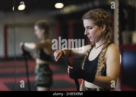 Eine junge Frau mit Zöpfen bereitet sich auf ein Training vor, sie wickelt einen engen Verband um ihre Hände, um sie zu schützen.das Fitness-Boxen-Workout der Frauen im Fitnessstudio Stockfoto