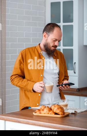 Junger Mann trinkt Kaffee Cappuccino mit Croissants und verwenden Smartphone in der Küche in gemütlichen Zuhause Interieur. Lächelnder kaukasischer 30s Glatze Mann haben Stockfoto