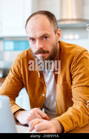 Portrait ruhiger Mann, der am Laptop in der Küche zuhause arbeitet. Kahlbärtiger 30s Mann, der die Kamera ansah Stockfoto