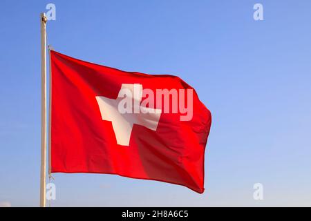 Die im Wind fliegende Schweizer Flagge wird an einem sonnigen Tag vor dem Hintergrund des blauen Himmels gesehen. Stockfoto