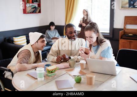 Junge Frau, die mit einem Tablet-pc am Tisch sitzt, mit einem Laptop und ihren Freunden im Zimmer Stockfoto