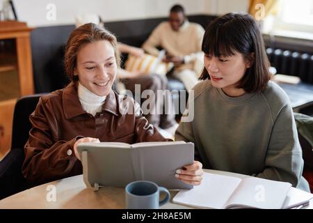 Zwei junge Frauen, die am Tisch sitzen und zusammen ein Buch lesen, bereiten sich auf die Hausaufgaben vor Stockfoto