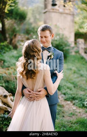 Bräutigam umarmt Braut Taille vor dem Hintergrund der alten Kapelle im Garten Stockfoto
