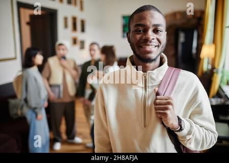 Porträt eines glücklichen afrikanischen Mannes mit Rucksack hinter seinem Rücken, der während des Standens an der Universität die Kamera anlächelt Stockfoto