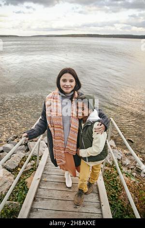 Glückliche junge kaukasische Frau und ihr süßer interrassischer Sohn in warmer Casualwear, die am Herbstwochenende gegen den See steht Stockfoto