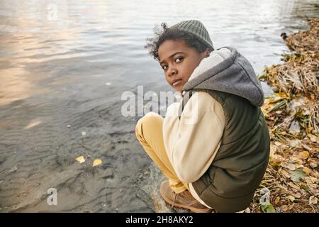 Süßer jugendlicher Junge in warmer Casualwear sitzt auf Kniebeugen am Wasser während der Kälte am Herbsttag und schaut dich an Stockfoto