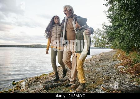 Ein glücklicher älterer Mann umarmt seine beiden Enkelkinder, während er sich am Wasser entlang bewegt und mit ihnen plaudert Stockfoto