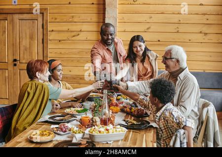 Fröhliche, moderne, interracial Familie, die mit Getränken über dem servierten Tisch klimmelte, während sie ihren Urlaub in häuslicher Umgebung feierte Stockfoto