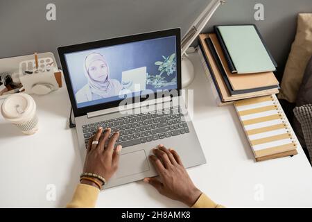 Laptop auf dem Schreibtisch mit Lehrer in Hijab auf dem Bildschirm während des Online-Unterrichts und Händen von afrikanischen männlichen Studenten über Tastatur Stockfoto