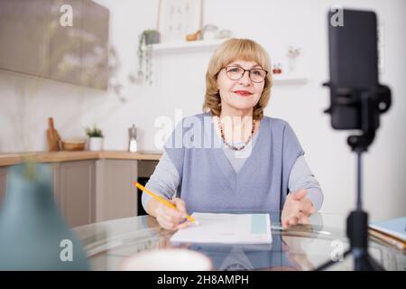 Blonde reife Frau in einer Brille spricht während des Livestreams oder Online-Trainings vor der Smartphone-Kamera Stockfoto