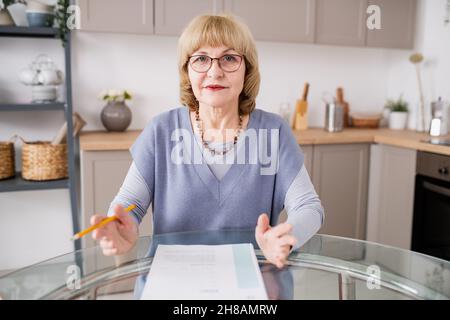 Blonde reife Lehrerin in Casualwear und Brillen, die den Schülern während des Online-Unterrichts etwas erklärt Stockfoto