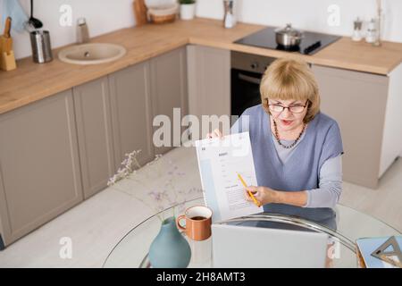 Blonde, selbstbewusste, reife Frau zeigt auf ein Finanzdokument und erklärt es dem Publikum während des Online-Unterrichts Stockfoto