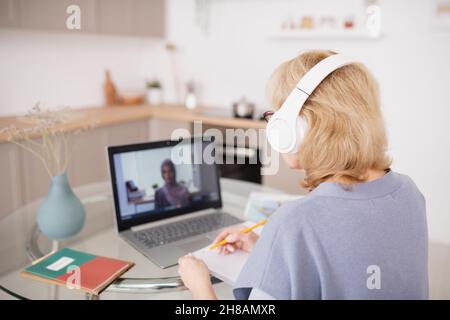 Rückansicht einer reifen Schülerin, die sich vor dem Laptop Notizen macht, während der Lehrer auf dem Bildschirm das neue Thema erklärt Stockfoto