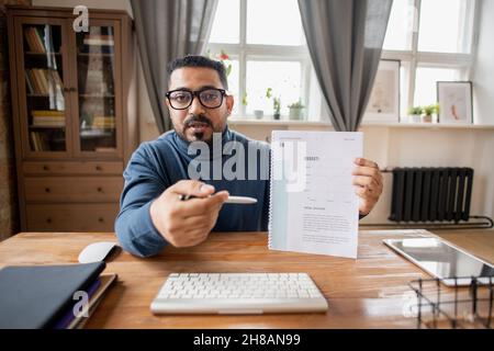 Selbstbewusster Wirtschaftslehrer, der mit einem Budgetdiagramm auf Finanzdokumente zeigt, während er vor dem Computer sitzt Stockfoto