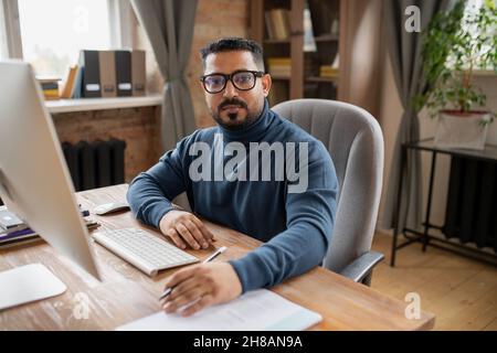 Ein ernsthafter Lehrer mittleren Alters, der in der häuslichen Umgebung am Schreibtisch vor dem Computermonitor die Kamera anschaut Stockfoto