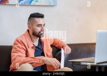 Junger, ernsthafter Mann, der während des Online-Unterrichts auf dem Sofa in der häuslichen Umgebung auf den Laptop-Bildschirm schaut Stockfoto