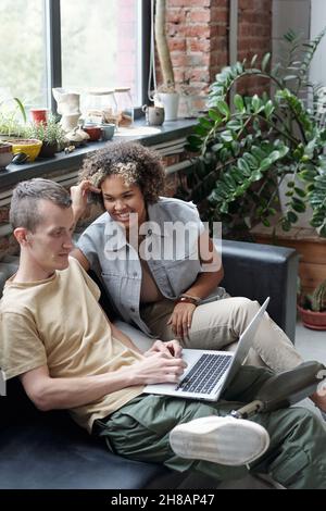 Glückliche junge interkulturelle Frau, die bei der Besprechung der Arbeitspunkte einen männlichen Kollegen mit Laptop ansieht Stockfoto