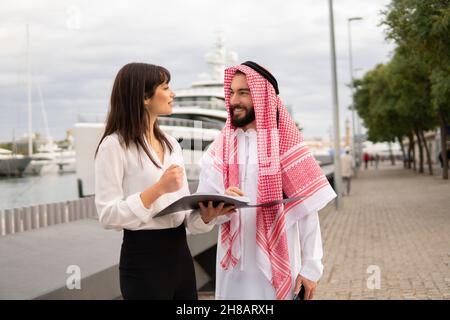 Junge europäische Geschäftsfrau und lächelnder arabischer Mandant in traditioneller Kleidung diskutieren Vertragsbedingungen und sprechen angenehm während des Treffens im Hafen, saudischer Geschäftsmann liest Dokumente vor der Unterzeichnung Stockfoto