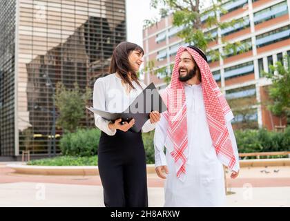 Verschiedene Geschäftsleute, arabischer Mann und kaukasische Frau, die Dokumente diskutieren, während sie auf der Stadtstraße spazieren gehen, junge lächelnde Geschäftsfrau und arabischer Mann, der bei einem Treffen im Freien einen Vertrag aushandelte Stockfoto