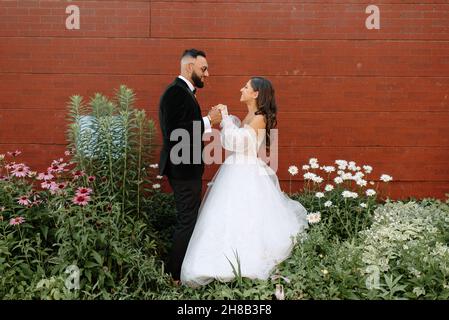 Braut und Bräutigam halten die Hände an Ziegelmauer und Blumen Stockfoto