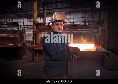 Schmiedearbeiter, zweiter Mitarbeiter, der die Hammersteuerung oder „Stampfer“ im vorformenden Schmiedebereich mit einer Reihe von Industriezangen befehligen kann, wenn die Ofentür geschlossen wird Stockfoto