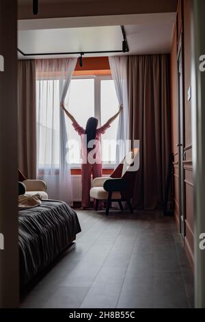 Rückansicht Morgenfrau im Schlafanzug Vorhänge öffnen Blick auf Fenster genießen neuen Tag im Schlafzimmer Stockfoto