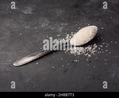 Granulierter weißer Zucker, auch Tischzucker in einem Löffel genannt, rustikaler dunkler Hintergrund, monochromatisches Thema mit Kopierraum Stockfoto