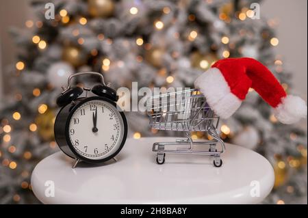 Weihnachtsgeschenke Zeit zum Einkaufen. Ein Einkaufswagen mit Weihnachtsmütze und Wecker am Weihnachtsbaum. Stockfoto