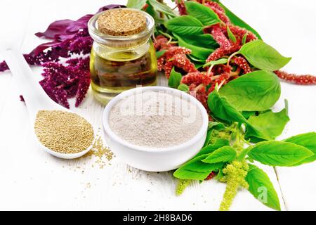 Amaranth-Mehl in einer Schüssel, Samen in einem Löffel und Öl in einem Glas, braune, grüne und violette Blüten einer Pflanze auf dem Hintergrund von hellem Holzbrett Stockfoto