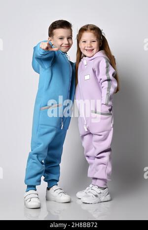 Zwei niedliche, lustige Kinder in Jumpsuits stehen von Angesicht zu Angesicht und der Junge zeigt mit dem Finger auf die Kamera Stockfoto