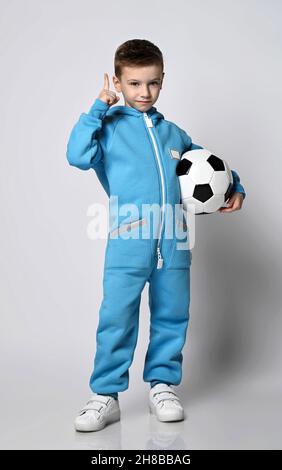 Frolic Kid Boy in blauem Jumpsuit mit Taschen steht mit Fußball in der Hand und deute den Zeigefinger nach oben Stockfoto