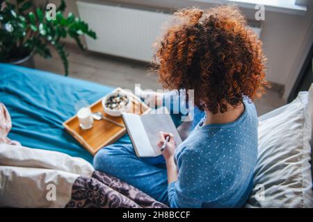 Rückansicht einer lockigen Frau, die auf dem Bett sitzt und etwas in seinem Notizbuch notiert. Tablett mit leckerem Frühstück auf dem Bett. Stockfoto