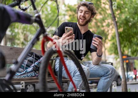 Ein hübscher Hipster-Typ trinkt Kaffee zum Mitnehmen auf einer Bank sitzend und macht einen Smartphone-Videoanruf - gen z People Habits Concept Stockfoto