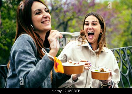Frauen, die Spaß haben, Essen zum Mitnehmen im Freien zu essen, sitzen auf einer Bank im Park mit Gesichtsmaske am Handgelenk. Neues normales Konzept der Begegnung junger Menschen Stockfoto