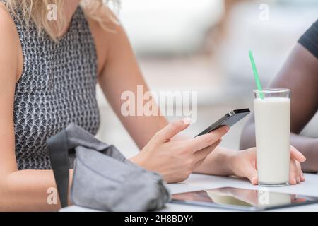 Zugeschnittenes Bild einer Person, die ihr Telefon benutzt und ein Glas Milchshake in einem Café im Freien hält Stockfoto