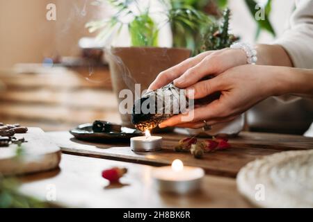 Frau Hände brennenden weißen Salbei, palo santo vor Ritual auf dem Tisch mit Kerzen und grünen Pflanzen. Rauch von Verschmierungen behandelt Schmerzen und Stress, klar Stockfoto