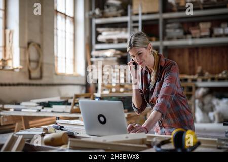 Frau mittleren Erwachsenen mit Laptop und Smartphone in Innenräumen in der Schreinerei, Konzept für kleine Unternehmen. Stockfoto