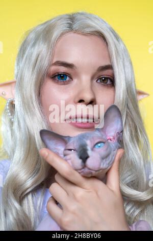 Porträt einer jungen blonden Frau cosplay Elf in blauem Kleid, hält Sphinx Katze, lächelnd, Blick auf die Kamera. Elf lockiges langes Haar, Augen verschiedener Farbe Stockfoto